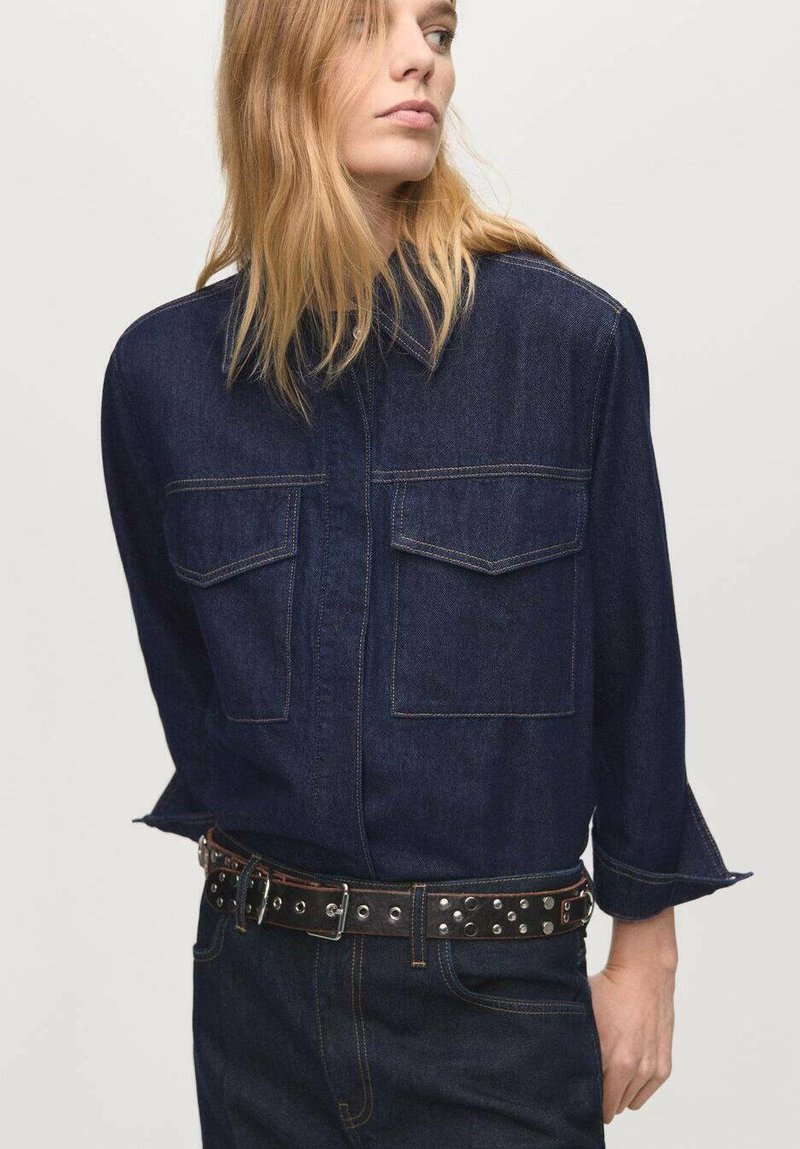 Camicia di jeans blu scuro con maniche lunghe, due tasche frontali e una patta con bottoni. Abbinata a una cintura nera con borchie argentate.
