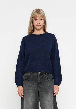 Marineblå strikkegenser med rund hals og lange, vide ermer. Kombinert med høyt midje svarte jeans med en falmet tekstur.