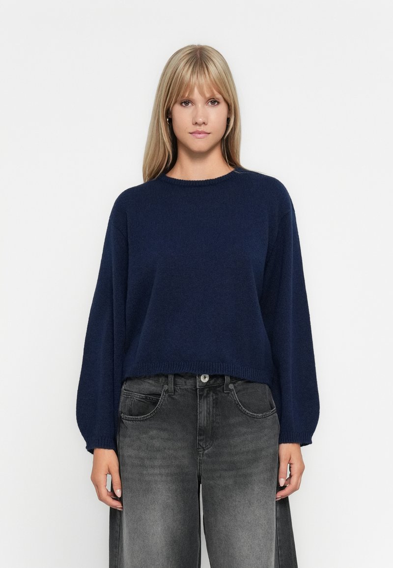 Marineblauw gebreid trui met een ronde hals en lange, brede mouwen. Gecombineerd met hoge zwarte jeans met een vervaagd textuur.