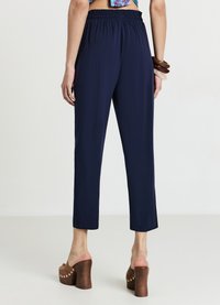 Pantalon à taille élastique bleu marine, longueur raccourcie, texture lisse. Modèle portant des mules marron à talons blocs, avec un top court à motifs et des bracelets.
