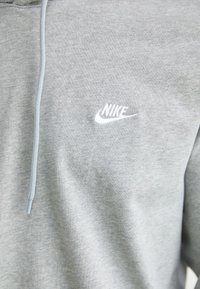 Siva bombažna trenirka s kapuco na vrvico, ki ima majhen bel Nike logotip na prsih. Mehka tekstura, sproščen dizajn.