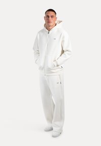 Sudadera blanca con cremallera y bolsillo frontal, combinada con pantalones de chándal blancos. Textura suave, diseño minimalista, con sutiles acentos de marca.