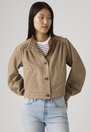 Žena s dlhými čiernymi vlasmi nosiaca béžový zapínaný kabátik cez bielo-čierno pruhované tričko a svetlomodré džínsy, pozerajúca sa doprava.