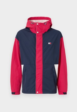 Veste coupe-vent colorée avec des panneaux bleu marine, rouge et blanc, une capuche avec cordon de serrage et des poches latérales. Matériau léger avec fermeture à boutons-pression.