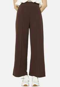 Pantalons amples marron en tissu doux, avec une taille haute, deux plis à l'avant, portés avec des baskets blanches.