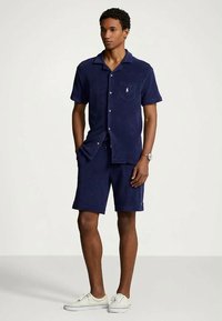 Man i marinblå kortärmad skjorta med knäppning och matchande shorts, iförd vita sneakers, står med händerna i fickorna.