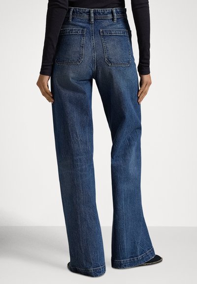 Άτομο που φοράει σκούρες μπλε τζιν παντελόνες με φαρδιά μπατζάκια και μαύρο μπλουζάκι με μακριά μανίκια, στέκεται με χαλαρά τα χέρια στα πλάγια μπροστά σε απλό φόντο.