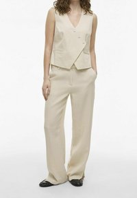 Beige, ilmanhihaista kietaisuliiviä, jossa on kaksi nappia ja sivutaskut, on yhdistetty löysästi istuviin beigeihin housuihin. Mustat tasapohjaiset kengät täydentävät asua.