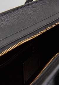 Musta nahkalaukku, jossa on teksturoitu pinta, kultainen vetoketjuyksityiskohta ja sileä, tumma vuori. Sisältää näkyvän merkkilapun.