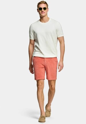 Junger Mann mit grüner Sonnenbrille, weißem Kurzarmhemd, korallenroten Shorts und beigen Loafers, der vor weißem Hintergrund nach vorne geht.