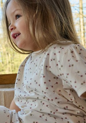 Tout-petit aux cheveux châtain clair portant une chemise blanche à petit motif floral, souriant et regardant vers la gauche à la lumière naturelle.
