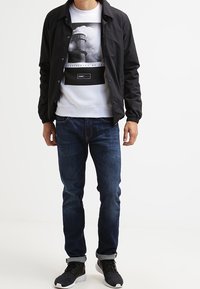 Schwarze leichte Jacke, weißer Pullover mit Grafikdruck, blaue Denim-Jeans und schwarze Sneakers. Lässiger Schnitt mit elastischen Bündchen.