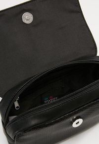 Černá texturovaná crossbody taška s klopou a stříbrným magnetickým zapínáním, částečně otevřená, aby odhalila vnitřní zipovou kapsu a textilní podšívku.