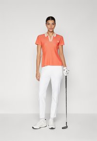 Πορτοκαλί κοντόμάνικο πόλο μπλουζάκι με λευκό γιακά, συνδυασμένο με λευκά παντελόνια και λευκά αθλητικά παπούτσια. Κράτημα γκολφ στο δεξί χέρι.