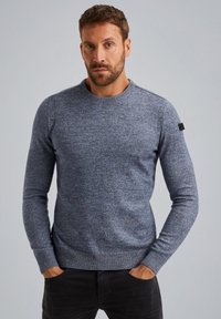 Maglione blu a collo giro con un motivo testurizzato, polsini e orlo a costine, e un piccolo etichetta nera sulla spalla sinistra. Indossato con pantaloni scuri.