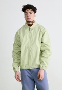 Jachetă fermoar de culoare verde deschis, cu guler și manșete elastice, având un logo mic pe piept, purtată cu pantaloni albaștri deschiși.