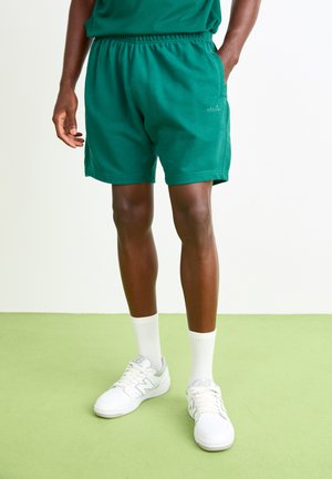 Green cotton shorts with an elastic waistband, side pockets, and a small logo on the front. Paired with white sneakers and socks.