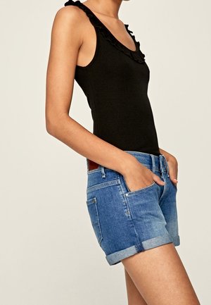 Woman wearing sleeveless black top with ruffled neckline and blue rolled-up denim shorts, hands in front pockets, neutral background.