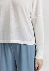 Witte, lichte gebreide top met lange mouwen, voorzien van een textuurpatroon en een ronde zoom. Gecombineerd met blauwe, losse broek.