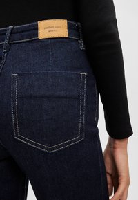 Sötétkék farmernadrág, magas derékkal, hátul világosbarna bőr címkével, kontrasztos varrással és két hátsó zsebből áll.