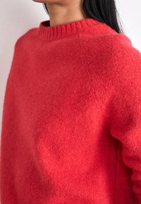 Czerwony sweter wykonany z miękkiego, puszystego materiału. Posiada prążkowany dekolt i dopasowany fason, gładką teksturę oraz minimalistyczne detale.