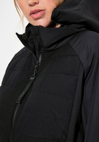 Chaqueta negra aislante con un alto cuello y capucha, con un patrón de acolchado texturizado y cierre de cremallera. La tela suave resalta el diseño.