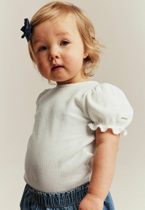 Kleinkind mit kurzen blonden Haaren und dunkelblauer Haarspange, trägt weißes Oberteil mit Puffärmeln und blaue Hosen mit geraffter Taille, schaut leicht zur Seite.