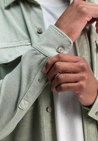 Lichtgroene button-up shirt gemaakt van zachte stof, met een borstzak, knoopmanchetten en een kraag. gedragen over een wit t-shirt.