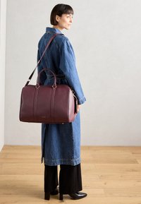 Burgundy leather duffel bag with dual handles and detachable shoulder strap, featuring smooth texture and subtle vertical seams.