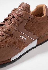 Baskets en cuir marron et daim avec des panneaux texturés, une bande blanche contrastante et un détail de logo sur le côté. Semelle en caoutchouc pour une meilleure adhérence.