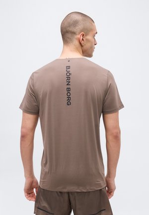 Mann mit rasiertem Kopf, trägt ein braunes kurzärmeliges Hemd mit vertikalem "BJÖRN BORG"-Schriftzug auf dem Rücken und passende Shorts, steht mit dem Rücken zur Kamera.