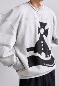 Grå överdimensionerad sweatshirt med svart grafisk design, med texturerad tryck och rundad halsringning. Modellen bär den med svarta byxor.