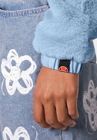 Sztuczne futro, jasnoniebieski rękaw z ciemnoniebieskim mankietem; dżinsy z dużymi białymi kwiatowymi wzorami. Czarna metka z pomarańczowym logo na mankiecie.