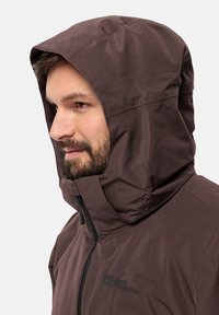 Giacca impermeabile marrone con cappuccio sagomato, chiusura con zip e dettaglio del logo. Presenta una texture liscia e un design aerodinamico.