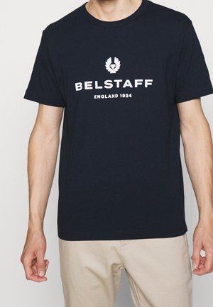 Mann trägt ein schwarzes Belstaff T-Shirt mit weißem Logo und Text "ENGLAND 1924", kombiniert mit beigen Hosen vor einfarbigem Hintergrund.
