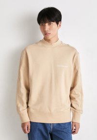 Felpa beige con collo alto, polsini e orlo a coste. Presenta un piccolo logo bianco sul lato sinistro del petto. Tessuto morbido, vestibilità comoda.