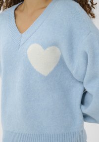 Pull tricoté bleu clair avec col en V. Présente une forme de cœur blanche douce sur la poitrine. Détails aux poignets et à l'ourlet en bord-côte.