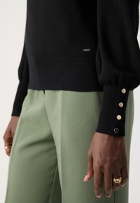 Schwarzer Strickpullover mit voluminösen Ärmeln und goldenen Knopfdetails an den Bündchen, kombiniert mit hoch taillierten dunkelgrünen Hosen.