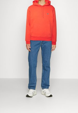 Person trägt ein leuchtend oranges Kapuzensweatshirt, blaue Jeans und weiße Turnschuhe und steht vor einem schlichten hellen Hintergrund.