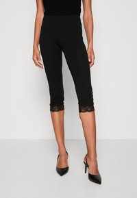 Leggings capri noirs en matière extensible, avec des accents en dentelle à l'ourlet. Assortis à des talons aiguilles noirs.