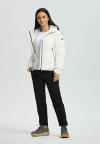 Kvinde står iført en hvid puffjakke, hvid skjorte, sorte bukser og brune vandresko med blå snørebånd mod en ensfarvet baggrund.