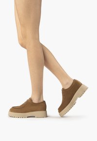 Scarpe in suede marrone con una spessa soletta beige, caratterizzate da una punta arrotondata e una minimali chiusura con lacci. Il design è casual e strutturato.