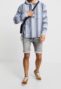 Sudadera con capucha a rayas en azul y gris con un bolsillo frontal, combinada con pantalones cortos de denim gris enrollados y zapatillas marrones. Mochila negra sencilla.