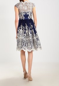 Marinblå klänning med spetsöverdrag, kortärmade och detaljerad blommig brodyr. Knä-lång fåll med scallopkant. Bärs med nakna klackar.