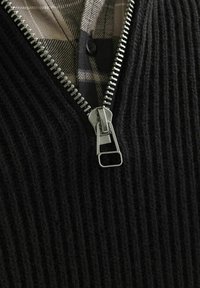 Schwarzer gerippter Pullover mit einem Metallreißverschluss am Hals, darunter ein karierter Hemd in Beigetönen und dunklen Farben.