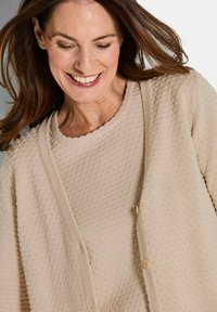 Beiger Cardigan mit strukturiertem Muster, Rundhalsausschnitt und Knopfverschluss. Weicher Stoff und lockere Passform. Dezente Details entlang der Kanten.