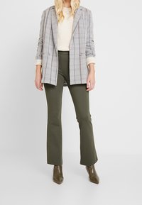 Grijze geruite blazer met twee knopen, crèmekleurige trui en olijfgroene flarebroek, gecombineerd met bruine hoge puntige laarzen.