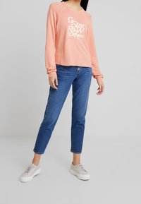 Sudadera de manga larga en melocotón claro con dobladillo sin rematar y logo en blanco de "GStar RAW Denim". Combinada con jeans de mezclilla azules y zapatillas gris claro.