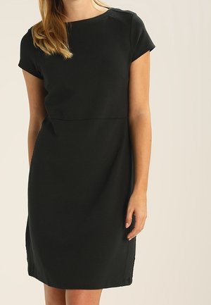 Donna che indossa un vestito nero a maniche corte, lungo fino al ginocchio, con scollatura rotonda, in piedi davanti a uno sfondo semplice.