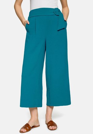 Femme portant un pantalon large court couleur sarcelle avec une ceinture assortie et des sandales marron, mains dans les poches.